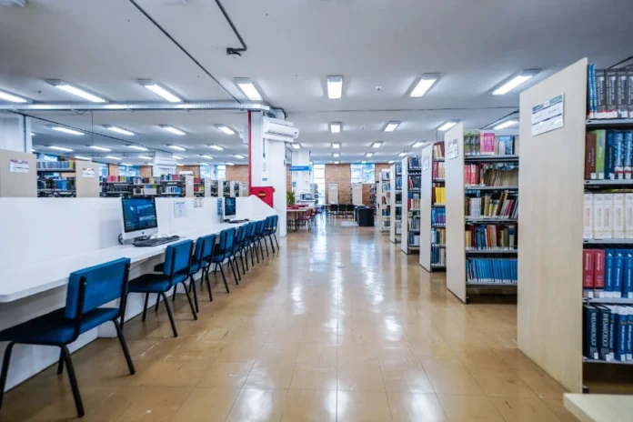 Alterações no Universidade Gratuita são aprovadas pela Alesc Uma ilustração de uma moderna biblioteca universitária, com estudantes engajados em leitura e pesquisa entre as estantes, simbolizando o acesso e a permanência no ensino superior promovidos pelo programa Universidade Gratuita.