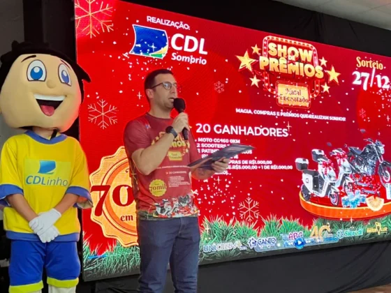 Mascote CDLinho e palestrante com microfone em frente ao painel de 'Show de Prêmios - Natal Encantado' da CDL Sombrio, no evento de capacitação em vendas.