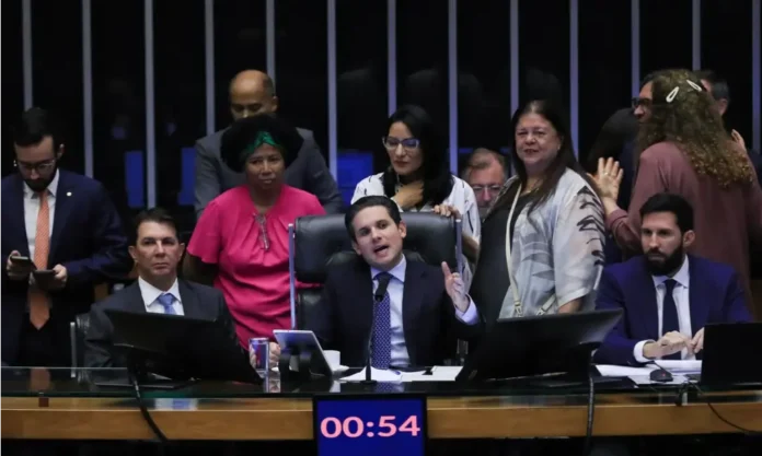 Câmara aprova licença de dois dias por sintomas menstruais Deputados e parlamentares em plenário da Câmara Federal, com um orador central gesticulando em frente a uma tela com a contagem de tempo, falando sobre a aprovação da licença menstrual.