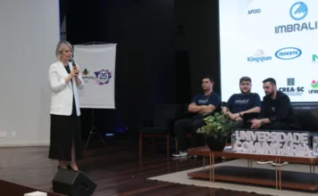 Construção 4.0: Simpósio da Unesc debate madeira e eficiência Reitora da UNESC discursando no palco durante o Simpósio Construção 4.0, onde se debate a eficiência e o uso da madeira na construção civil em Criciúma.