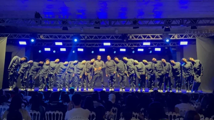 Coreógrafa da Unesc é destaque em festival com danças urbanas Grande grupo de dançarinos de danças urbanas em performance no palco, sob luzes azuis, representando o trabalho da coreógrafa da UNESC em festival.