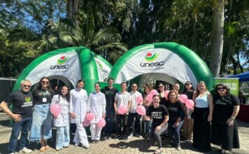 Criciúma: Unesc une Outubro Rosa e Dia das Crianças em praça Equipe da UNESC, incluindo estudantes de jaleco e voluntários, em frente a tendas infláveis verdes, segurando balões rosa em ação de conscientização do Outubro Rosa em uma praça de Criciúma.