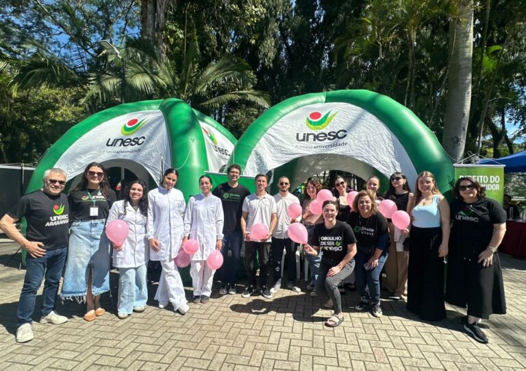 Criciúma Unesc une Outubro Rosa e Dia das Crianças em praça Equipe da UNESC, incluindo estudantes de jaleco e voluntários, em frente a tendas infláveis verdes, segurando balões rosa em ação de conscientização do Outubro Rosa em uma praça de Criciúma.