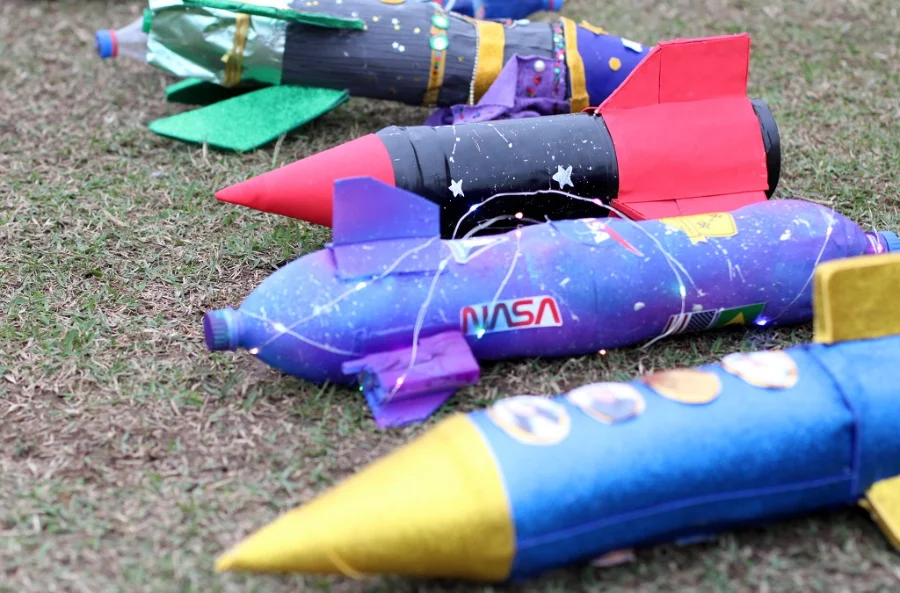 Foguetes artesanais feitos de garrafas PET, decorados com cores vibrantes e brilho, deitados na grama e prontos para a Competição Interclubes de Foguetes em Criciúma.