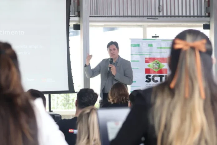 Criciúma sedia Ignition Startup e anuncia R$ 20 mi para crédito Palestrante gesticula em frente a uma plateia durante o evento Ignition Startup em Criciúma, com o banner da SCTI (Secretaria de Ciência, Tecnologia e Inovação) de Santa Catarina ao fundo.
