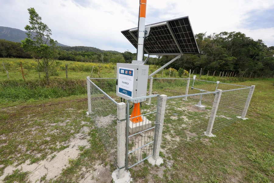 Estação hidrometeorológica automática, alimentada por painel solar e protegida por cerca, recém-instalada pela Defesa Civil para monitoramento de chuvas e rios em Santa Catarina.