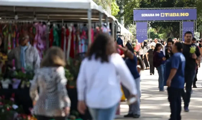 Desemprego cai a 5,6% e atinge menor taxa da série histórica Pessoas transitando em área externa de feira de empregos ou eventos ("Semana do Trabalho" visível), simbolizando a queda da taxa de desemprego no Brasil para 5,6%.