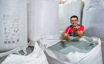 FIESC: Novo decreto de logística reversa eleva custo Empresário sorrindo, com as mãos em grânulos de plástico reciclado dentro de um grande saco, representando a indústria de Santa Catarina que debate o aumento de custos com o novo decreto de logística reversa.