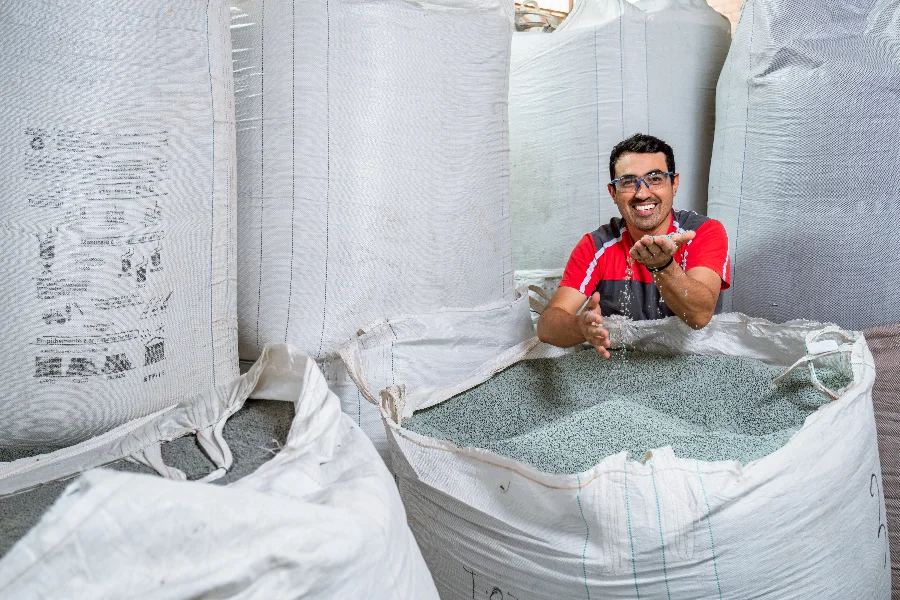 FIESC Novo decreto de logística reversa eleva custo Empresário sorrindo, com as mãos em grânulos de plástico reciclado dentro de um grande saco, representando a indústria de Santa Catarina que debate o aumento de custos com o novo decreto de logística reversa.
