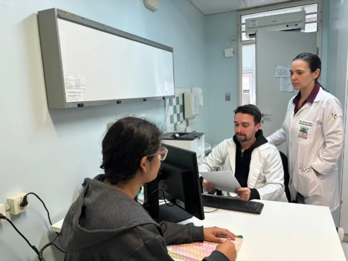 Faltas em hospitais do estado prejudicam o SUS e atrasam filas Dois profissionais de saúde (médicos) em jaleco atendendo uma paciente em consultório, ilustrando o impacto das faltas médicas que atrasam as filas do SUS em Santa Catarina.