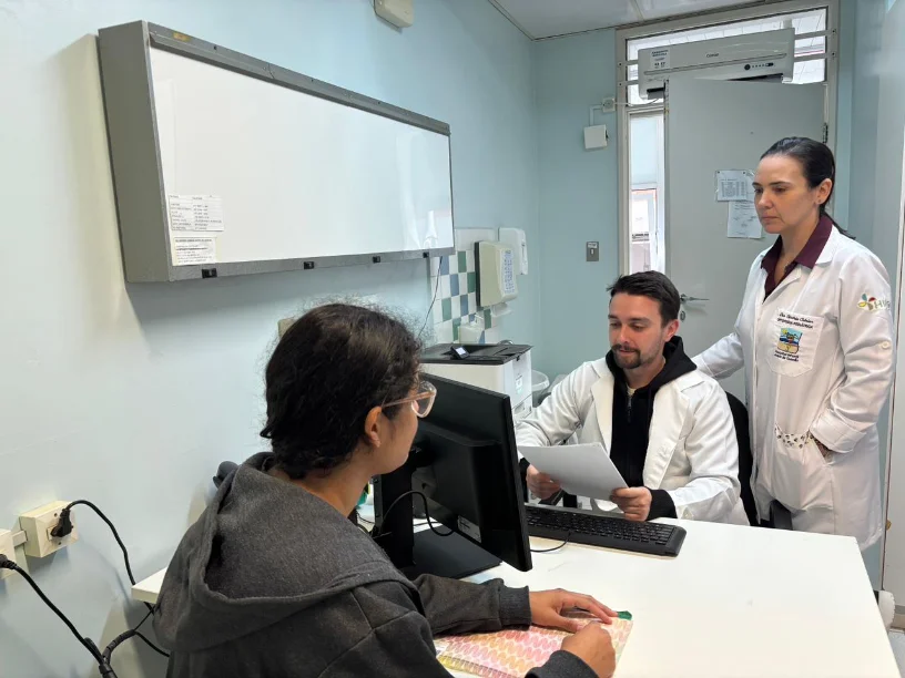 Faltas em hospitais do estado prejudicam o SUS e atrasam filas Dois profissionais de saúde (médicos) em jaleco atendendo uma paciente em consultório, ilustrando o impacto das faltas médicas que atrasam as filas do SUS em Santa Catarina.