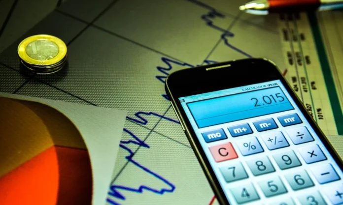Focus Mercado reduz projeção do IPCA pela terceira semana Calculadora em tela de celular, sobre gráficos e moedas, representando a redução da projeção do IPCA (inflação) pelo Boletim Focus do Banco Central.