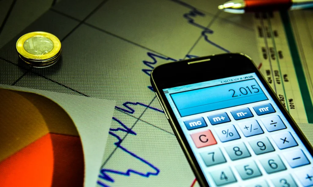 Focus Mercado reduz projeção do IPCA pela terceira semana Calculadora em tela de celular, sobre gráficos e moedas, representando a redução da projeção do IPCA (inflação) pelo Boletim Focus do Banco Central.