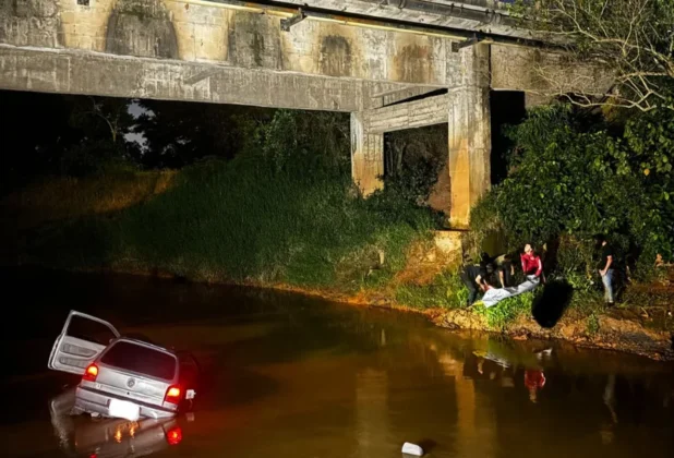 Carro parcialmente submerso em rio escuro, sob uma ponte de concreto, com equipes de resgate na margem após o acidente fatal em Forquilhinha.