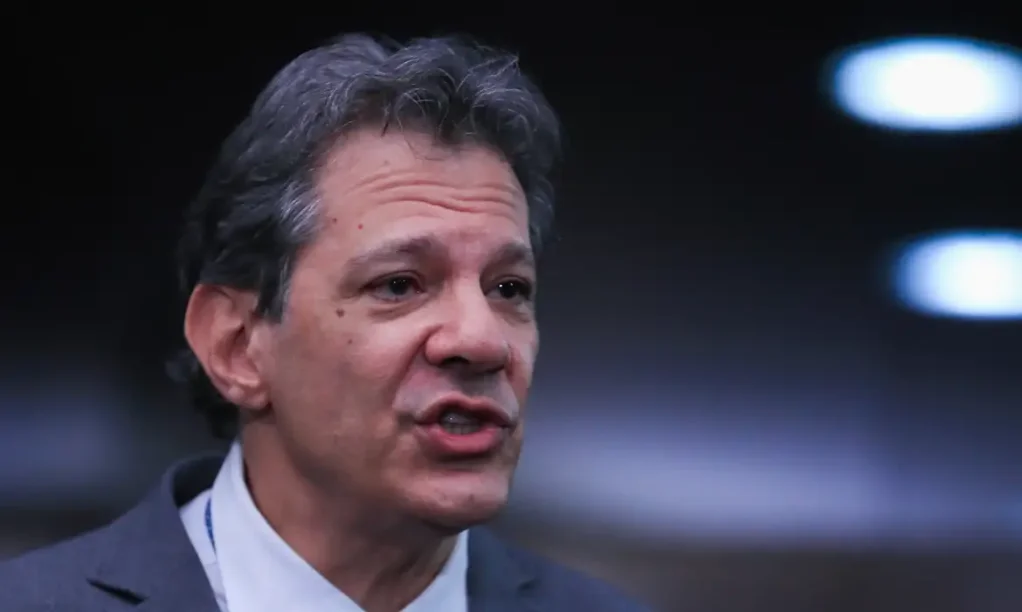 Haddad negocia compensação para isenção do Imposto de Renda Ministro Fernando Haddad, usando terno e gravata, falando em um evento ou entrevista em local interno com fundo escuro.