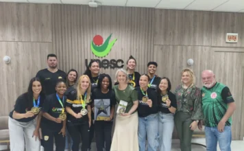 Handebol da Unesc é recebido após vaga em Mundial na França Equipe de Handebol Feminino da Unesc posa para foto com a reitora, segurando medalhas de ouro, em uma sala com o logotipo da universidade ao fundo.