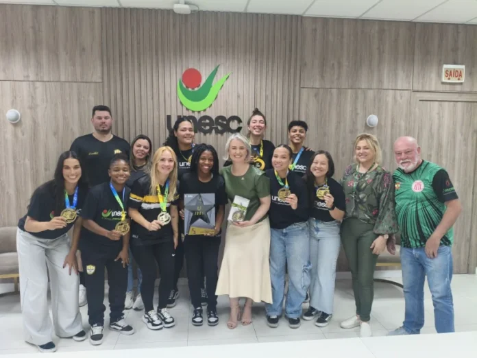 Handebol da Unesc é recebido após vaga em Mundial na França Equipe de Handebol Feminino da Unesc posa para foto com a reitora, segurando medalhas de ouro, em uma sala com o logotipo da universidade ao fundo.