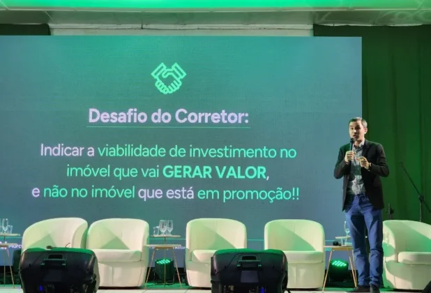 Palestrante Marcelo Velozo, em pé em frente a um telão verde com a frase "Desafio do Corretor" e cadeiras brancas, em Fórum Empresarial em Sombrio.