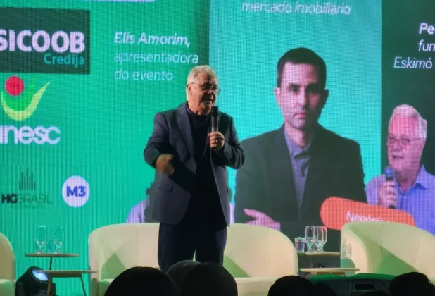 Close-up no palestrante Pedro Reis do IV Fórum Empresarial ACIS em Sombrio, falando ao microfone com imagens de outros convidados projetadas no telão.