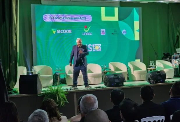 Palestrante Pedro Reis, discursando com microfone no centro do palco, com cadeiras e o logo da ACIS ao fundo, no Fórum Empresarial em Sombrio.
