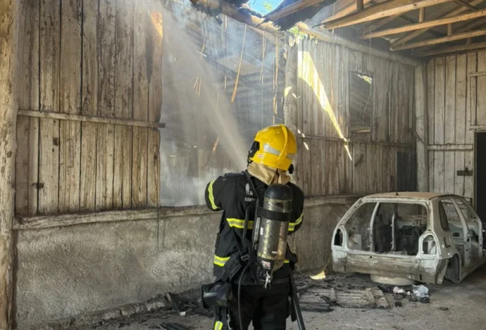 Incêndio atinge galpão abandonado em Criciúma Bombeiro com capacete amarelo combatendo um incêndio em um galpão abandonado de madeira, com o esqueleto de um carro queimado ao lado, em Criciúma.