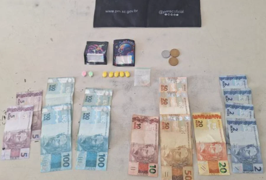 Jovem tenta fugir, mas é preso por tráfico em Arroio do Silva Material apreendido pela Polícia Militar em Arroio do Silva: diversas notas de Real (R$ 100, R$ 50, R$ 20, R$ 10, R$ 5, R$ 2) e diferentes tipos de drogas, incluindo pílulas e embalagens seladas, após prisão por tráfico.