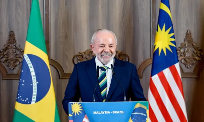 Lula e Trump criam canal direto para negociação Presidente Lula sorrindo, discursando em púlpito com a bandeira do Brasil e da Malásia, em evento diplomático sobre negociações com os Estados Unidos.
