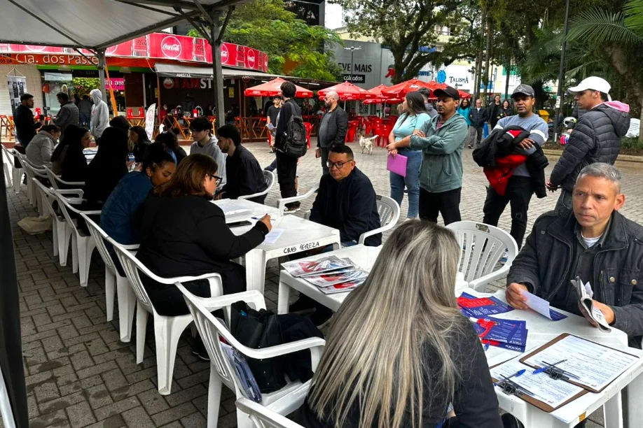 Candidatos em uma feira de empregos ao ar livre, sentados em mesas sendo atendidos por recrutadores no Balcão de Empregos.