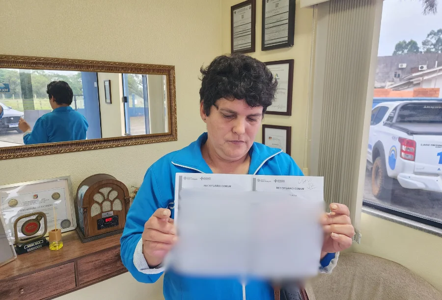 Moradora de Sombrio pede ajuda para comprar remédios Moradora de Sombrio segurando receitas médicas em frente a um balcão, ilustrando a necessidade de ajuda financeira para comprar remédios.