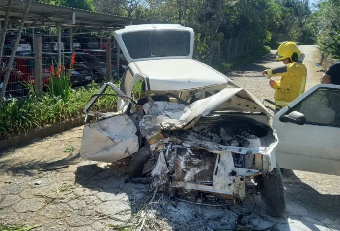 Motorista fica ferido após Uno colidir com caminhão em Içara Carro Fiat Uno branco com a frente completamente destruída após colidir com caminhão em Içara, com socorrista de capacete amarelo prestando atendimento no local.