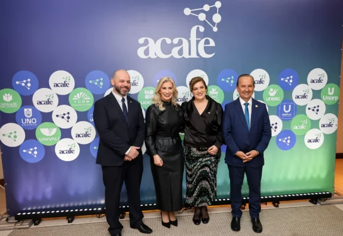 Nova presidente da Acafe destaca Universidade Gratuita Nova presidente da ACAFE e outras autoridades em frente ao logo da instituição, destacando o programa Universidade Gratuita e a importância da educação em Santa Catarina.