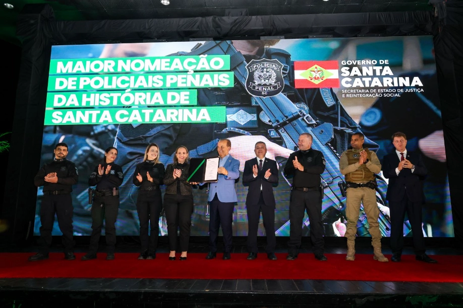 Polícia Penal de SC atinge maior efetivo da história Governador Jorginho Mello (ao centro) com a Secretária Danielle Amorim Silva e um grupo de novos Policiais Penais no palco, celebrando a maior nomeação da história de Santa Catarina.