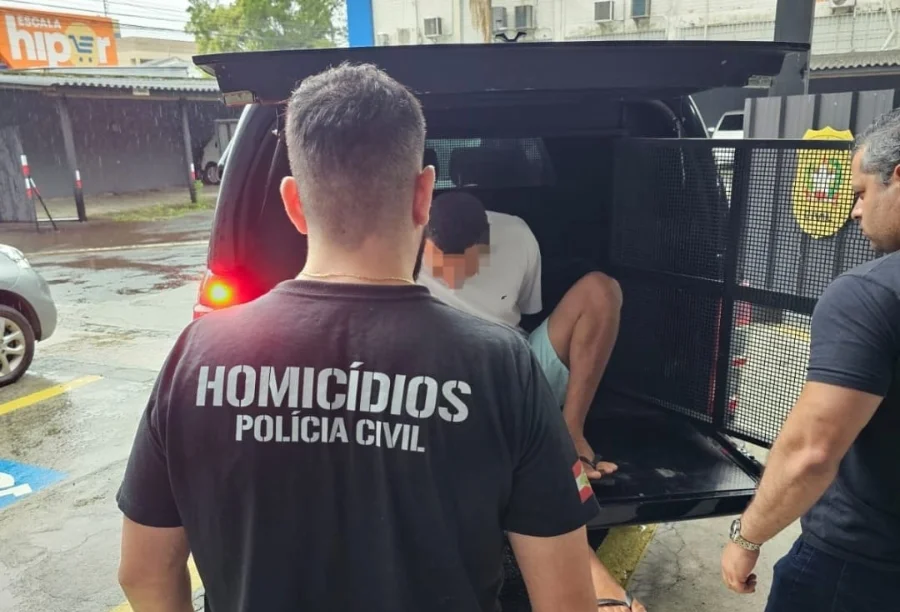 Homem de 21 anos, suspeito de tentativa de homicídio, sendo conduzido por policial à viatura da Polícia Civil em Criciúma.