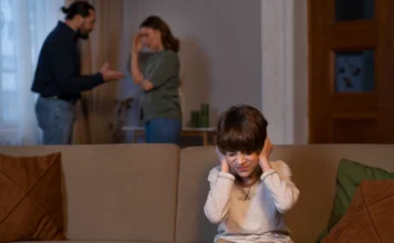 Quando o amor vira arma: os impactos da alienação parental Criança sentada em um sofá cobrindo os ouvidos com expressão de dor, com os pais discutindo ao fundo (desfocados), ilustrando o impacto emocional da alienação parental.
