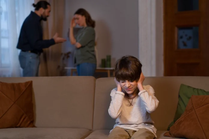 Quando o amor vira arma os impactos da alienação parental Criança sentada em um sofá cobrindo os ouvidos com expressão de dor, com os pais discutindo ao fundo (desfocados), ilustrando o impacto emocional da alienação parental.