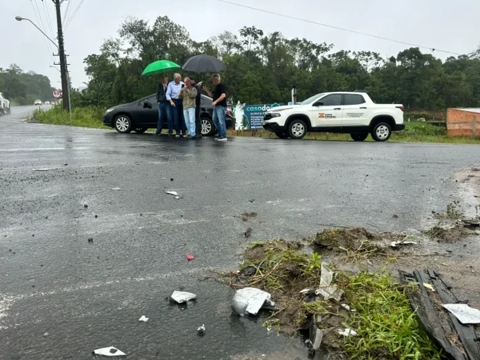 SC-442 Cocal do Sul pede rótulas e trevo para reduzir acidentes Autoridades de Cocal do Sul e técnicos reunidos na SC-442, sobre destroços de acidente na pista molhada, discutindo a necessidade de rótulas e trevo para aumentar a segurança.