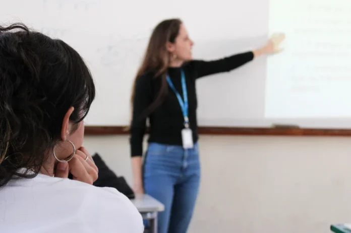 SED SC abre inscrição para seleção de professores ACTs Professora em sala de aula apontando para um quadro branco durante a aula, representando a seleção de professores ACTs da Secretaria de Educação de Santa Catarina (SED SC).