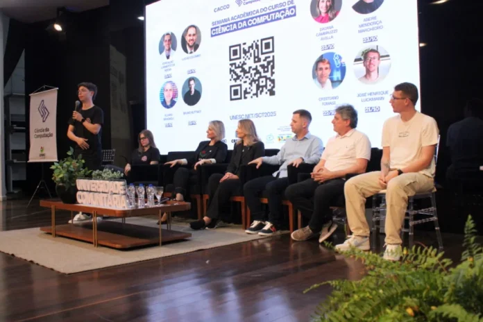 Semana de Computação da Unesc debate agentes inteligentes Mesa de debate no palco da UNESC (Criciúma) com palestrantes e apresentador, durante a Semana de Computação para discutir agentes inteligentes e Ciência da Computação.
