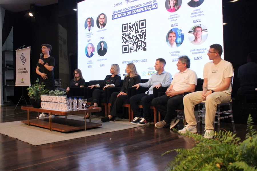 Semana de Computação da Unesc debate agentes inteligentes Mesa de debate no palco da UNESC (Criciúma) com palestrantes e apresentador, durante a Semana de Computação para discutir agentes inteligentes e Ciência da Computação.