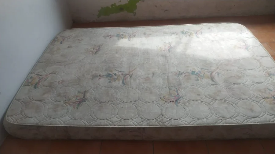 Colchão simples e antigo colocado diretamente no chão de uma casa com sinais de deterioração em Sombrio, ilustrando a situação de vulnerabilidade da família que precisa de doações.