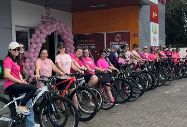 Mulheres sorrindo em suas bicicletas, em frente ao arco de balões rosa do Sombrio Moda Shopping, prontas para o "Pedal Rosas Sobre Rodas" em Sombrio.