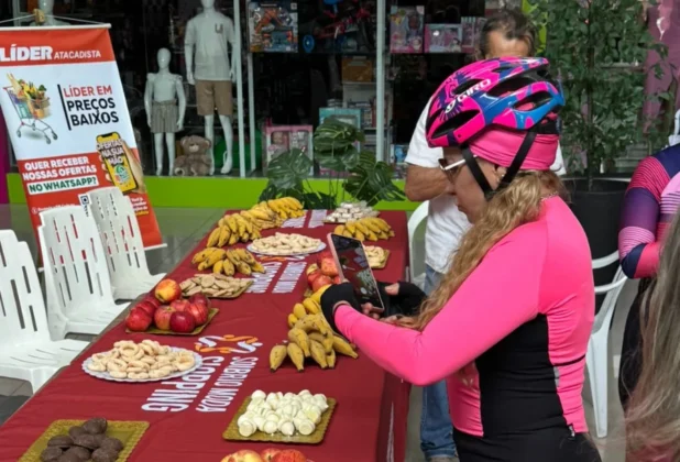 Ciclista com capacete rosa e luvas tira foto de mesa de frutas (maçãs e bananas) e biscoitos, oferecida como apoio nutricional no "Pedal Rosas Sobre Rodas" no Sombrio Moda Shopping.