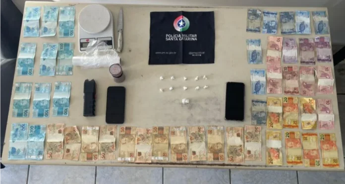 Três são presos por tráfico em flagrante pela PM de Araranguá Material apreendido em flagrante de tráfico em Araranguá: dinheiro em notas de Real, balança de precisão, celulares, entorpecentes e uma arma branca, dispostos sobre uma mesa.