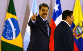 Trump anuncia captura de Nicolás Maduro em ataque militar O presidente da Venezuela, Nicolás Maduro, sorrindo e fazendo o sinal V de vitória com a mão, durante evento internacional, com bandeiras de países como Brasil e Chile ao fundo.
