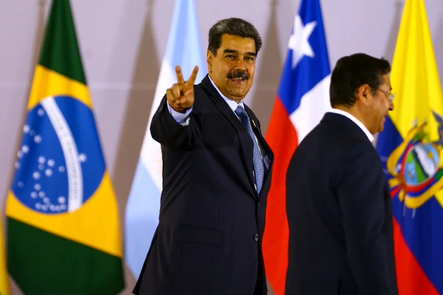 Trump autoriza CIA a realizar operações secretas na Venezuela O presidente da Venezuela, Nicolás Maduro, sorrindo e fazendo o sinal V de vitória com a mão, durante evento internacional, com bandeiras de países como Brasil e Chile ao fundo.