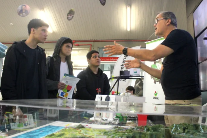 Unesc Profissões orienta 5,7 mil alunos do Terceirão na região Professor da UNESC gesticulando e orientando um grupo de alunos do Terceirão em um estande de profissões, em evento que atendeu 5,7 mil estudantes da região.