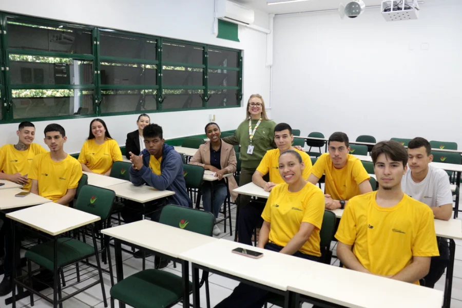 Jovens estudantes em sala de aula na UNESC (Criciúma) participam de palestra sobre o impacto das redes sociais e a ansiedade, com instrutora ao fundo.