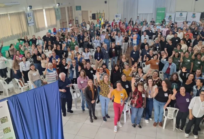 Unesc leva etnobotânica e plantas bioativas a seminário no RS Vista de cima de uma grande plateia em um seminário, com dezenas de participantes levantando as mãos e sorrindo, representando o evento sobre etnobotânica da Unesc.