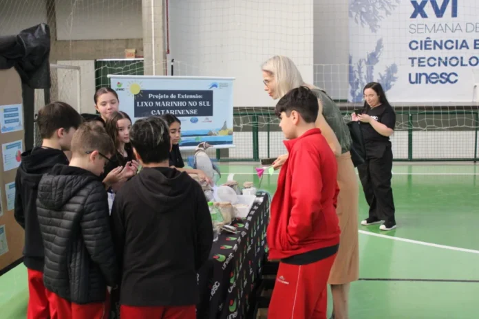 Unesc sedia feira com foco em ciência e educação básica Alunos da educação básica e professora em volta de um estande sobre 'Lixo Marinho', durante a Feira de Ciência e Tecnologia sediada pela UNESC em Criciúma.