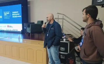 Amesc sedia 2º Fórum de Turismo Caminho dos Canyons Palestrante com microfone, em pé no palco, em frente ao painel do "Fórum de Turismo - Caminho dos Canyons", evento da AMESC realizado em Araranguá para debater a inovação no setor.